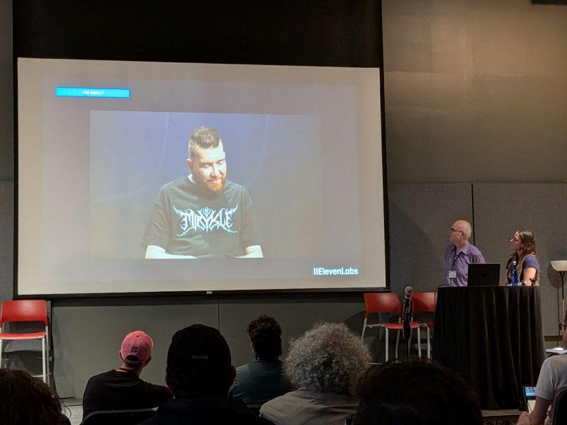 A conference screen labelled THE IMPACT shows a bearded musician in a black metal band shirt next to the ElevenLabs logo, with two presenters at a podium