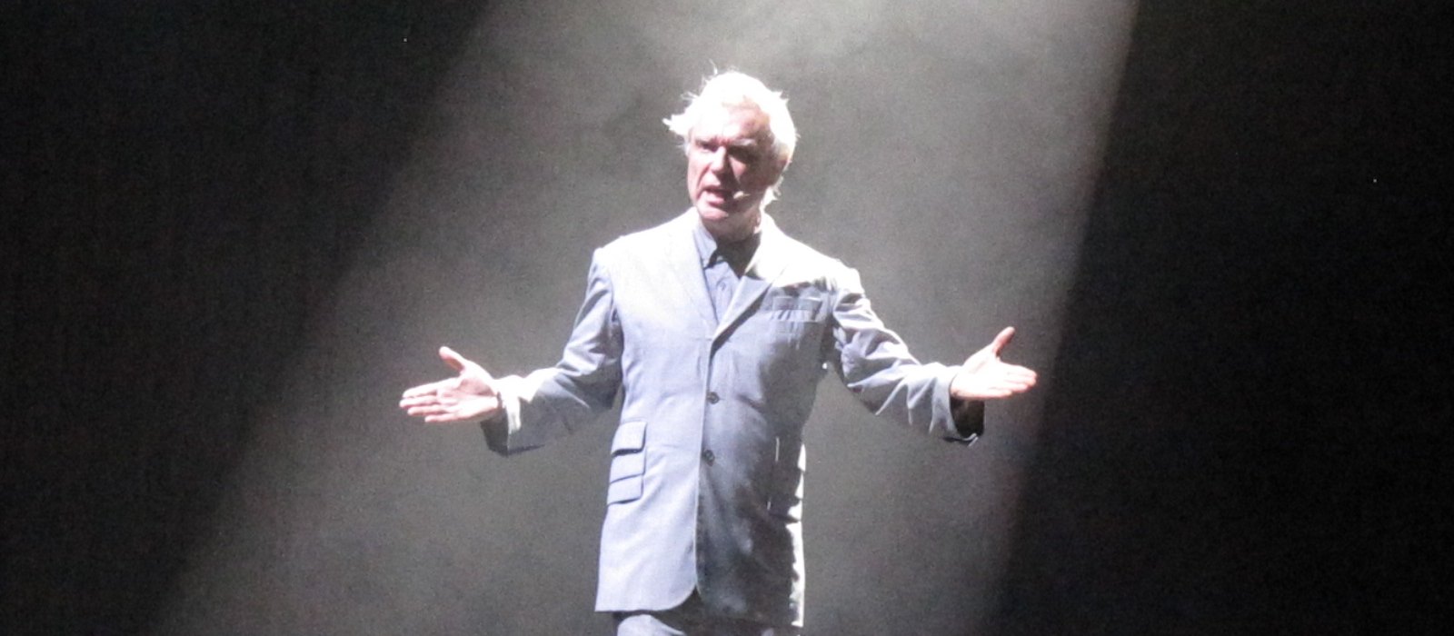 David Byrne in a grey suit with his arms open under a spotlight on stage in 2018