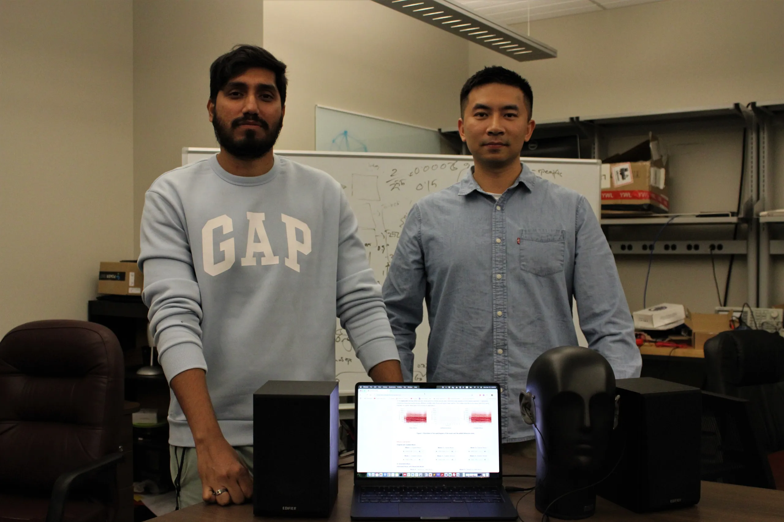 Two men standing in a lab setting with a table in front of them displaying a laptop, two speakers, and a mannequin head with headphones. The laptop shows audio analysis or AI tools on its screen, and a whiteboard with calculations is visible in the background.