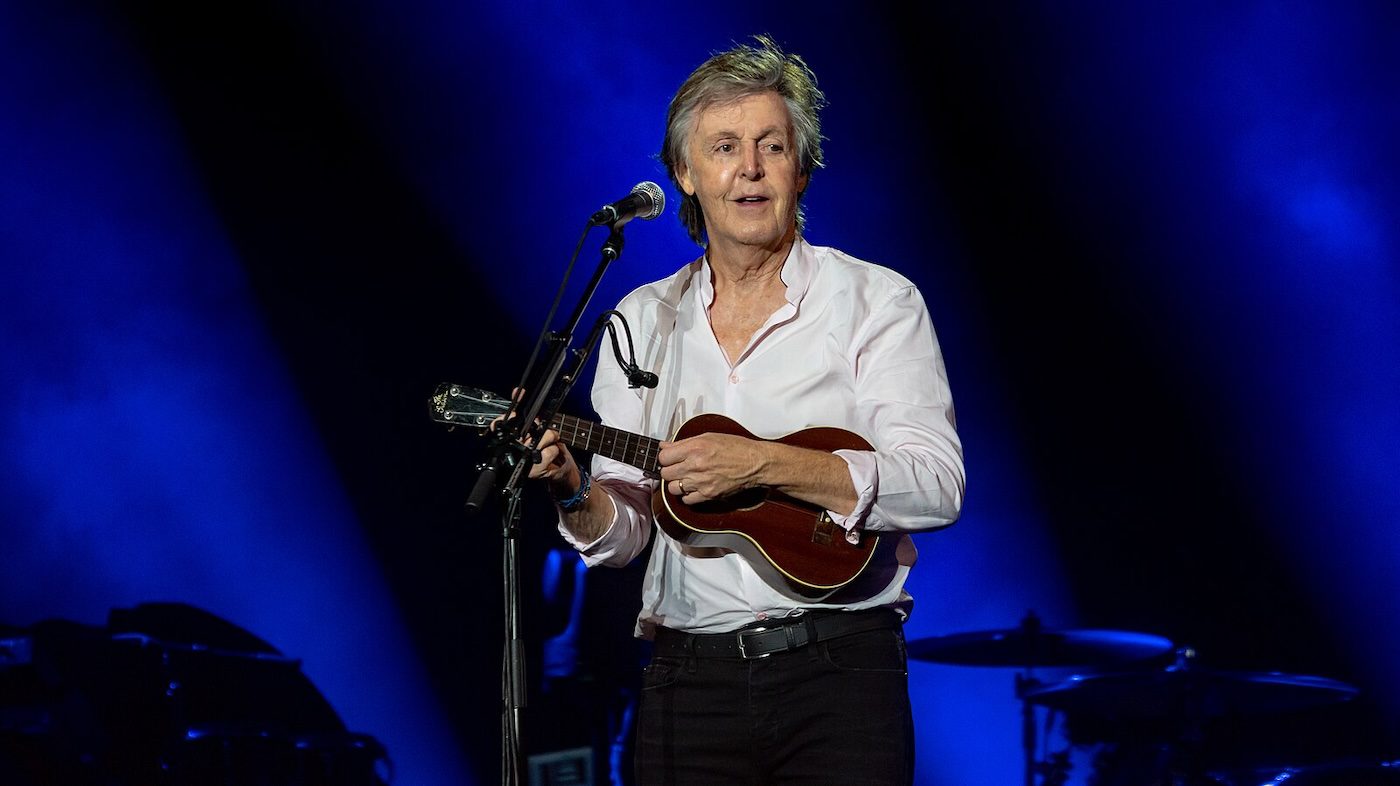 A live performance photo of Paul McCartney playing a small guitar on stage. He wears a white shirt and is illuminated by blue stage lighting with microphones set up around him.