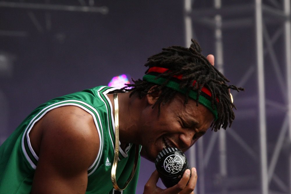 A live performance shot of Lupe Fiasco passionately singing into a microphone, wearing a green sports jersey and a red-and-green headband. The background shows stage equipment and a dimly lit environment.