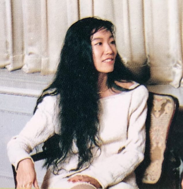 A vintage photo of Yumi Matsutoya sitting on a chair, wearing a white sweater and smiling.