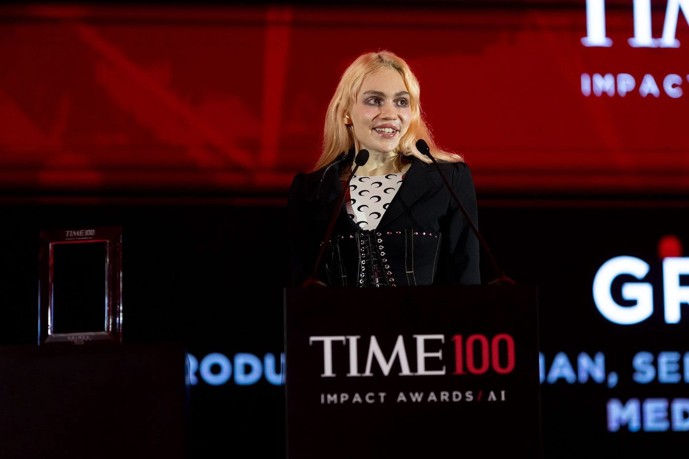 Grimes speaks at the TIME100 Impact Awards in Dubai, standing at a podium with a red-lit backdrop.