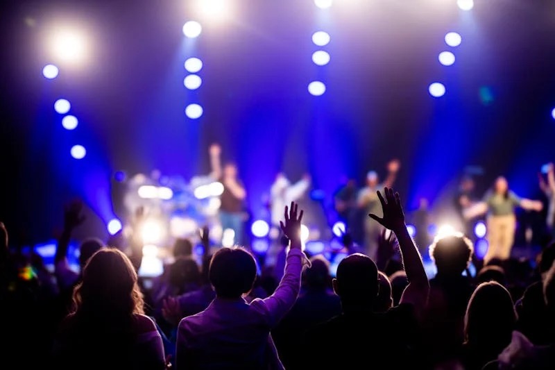 Crowd raising hands at a concert with bright blue stage lights and blurred performers on stage.